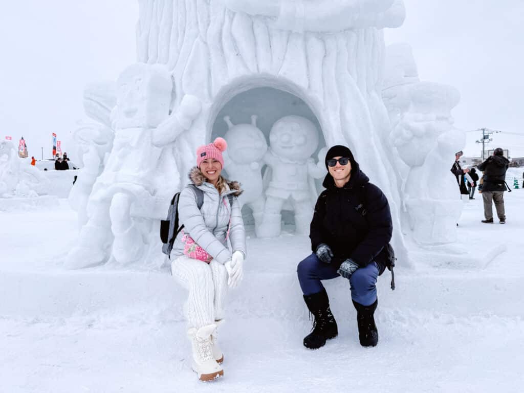 Couple in winter gear sitting on a snow bench in front of an Anpanman character snow sculpture at Asahikawa Winter Festival.