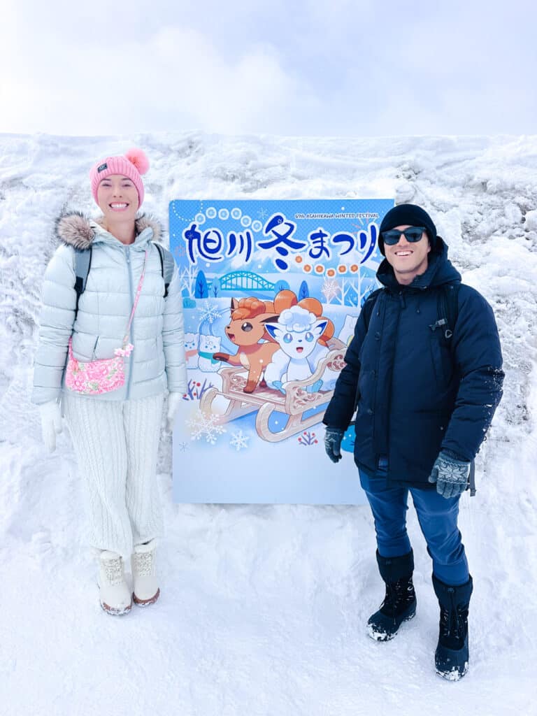 Couple posing in front of the official Asahikawa Winter Festival 2026 poster sign featuring Vulpix and Ponyta on a sled.