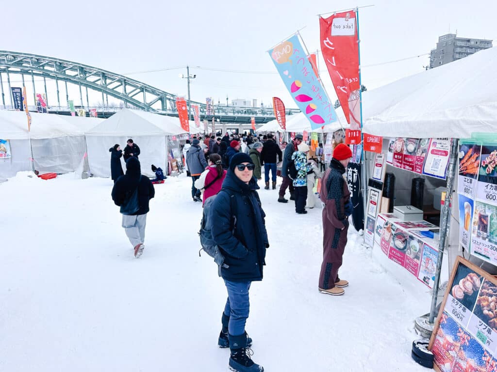 Festival food stalls and visitors gathered beside the green Asahibashi Bridge at Asahikawa Winter Festival, Hokkaido.