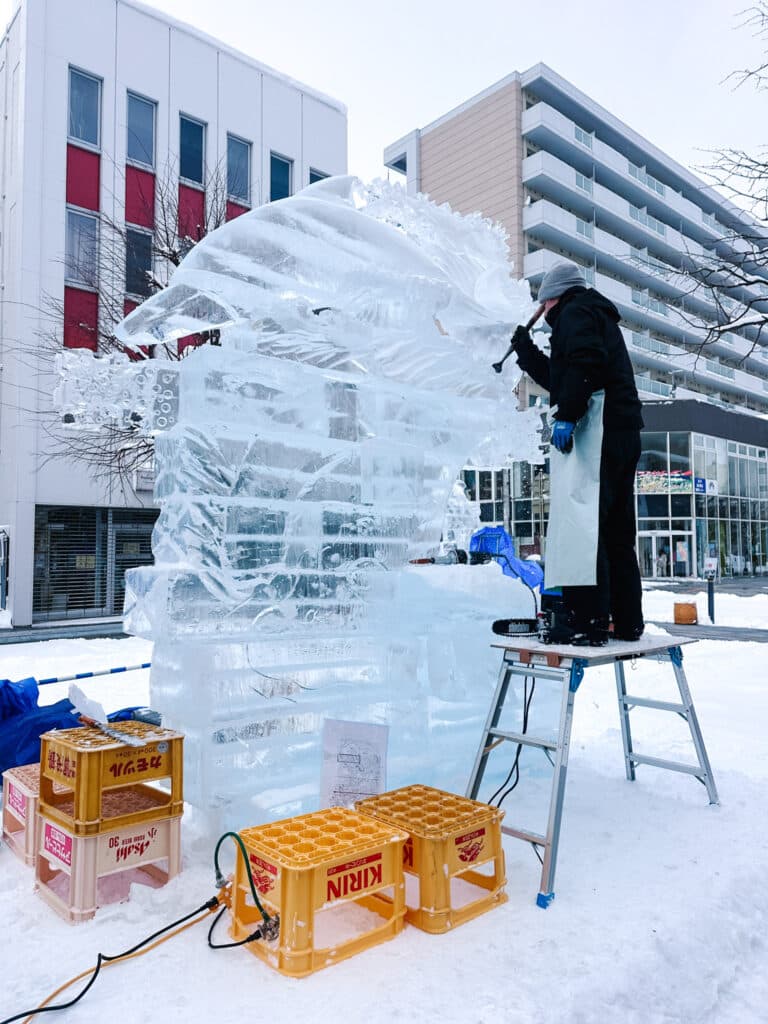 ce sculptor on a ladder carving a large fish-shaped sculpture in downtown Asahikawa, with Kirin and Asahi beer crates stacked at the base.