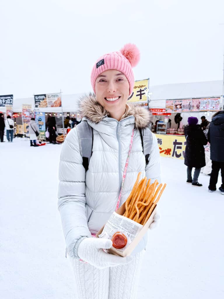 Close-up of long crispy Japanese potato fries with a cup of ketchup, held by a woman in white knit pants at Asahikawa Winter Festival.