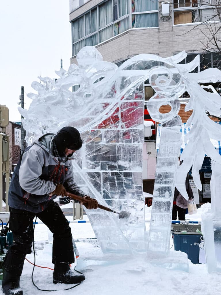 Ice artist in a grey hoodie chiselling intricate detail into a tall sculpture at Asahikawa Winter Festival.