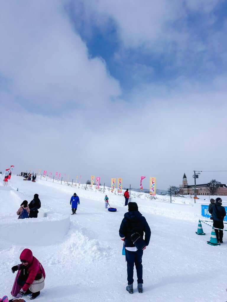 Visitors climbing the "Hankyu Slider" snow tubing hill lined with colourful Japanese flags at Asahikawa Winter Festival.