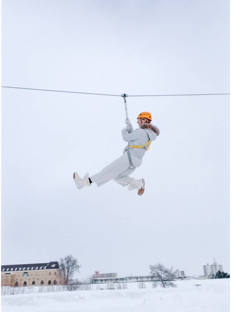 Woman in a white snowsuit and orange helmet ziplining mid-air above the snow at Asahikawa Winter Festival.