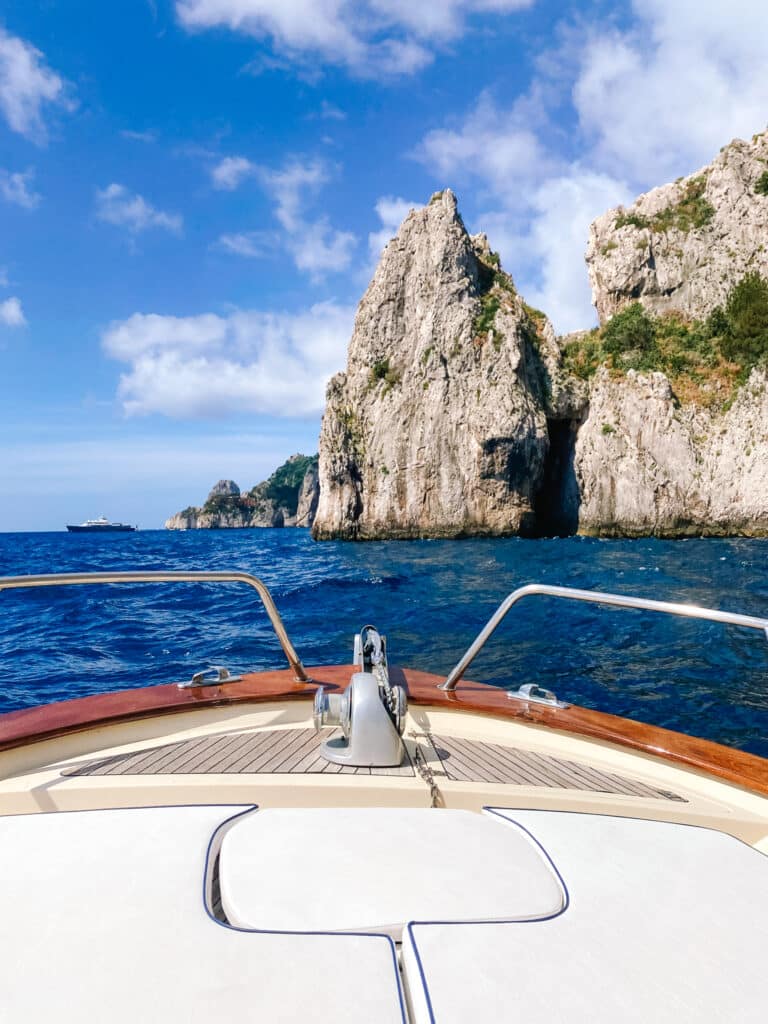 View from the bow of a wooden boat approaching the towering limestone cliffs and Faraglioni rocks of Capri, Italy
