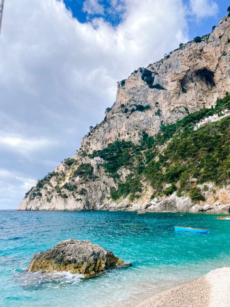 Turquoise cove water beneath dramatic Capri cliffs with a single blue rowing boat floating near the rocky shore of Marina Piccola on Capri