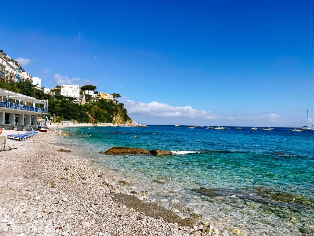 Sun loungers lined up at a beach club on Capri's pebble shore with turquoise water and boats moored offshore of Marina Grande