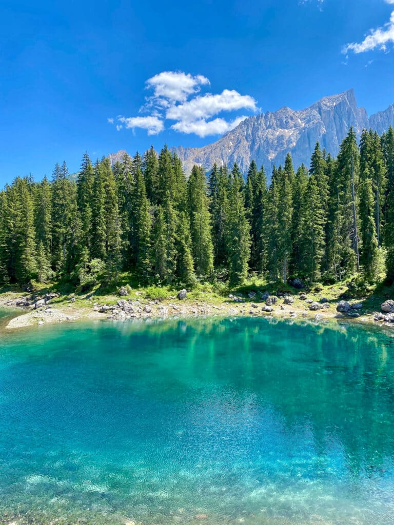 Portrait view of Lago di Carezza's (Karersee) clear turquoise water and pine-fringed shoreline with mountain peaks under a cloud-streaked sky