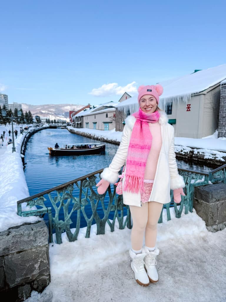 Woman in a white coat and pink scarf on the snowy Otaru Canal path, a cruise boat passing icicle-fringed warehouses