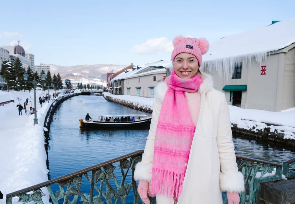 Woman at the Otaru Canal railing on a bright day, snow-covered warehouses and a cruise boat across the water