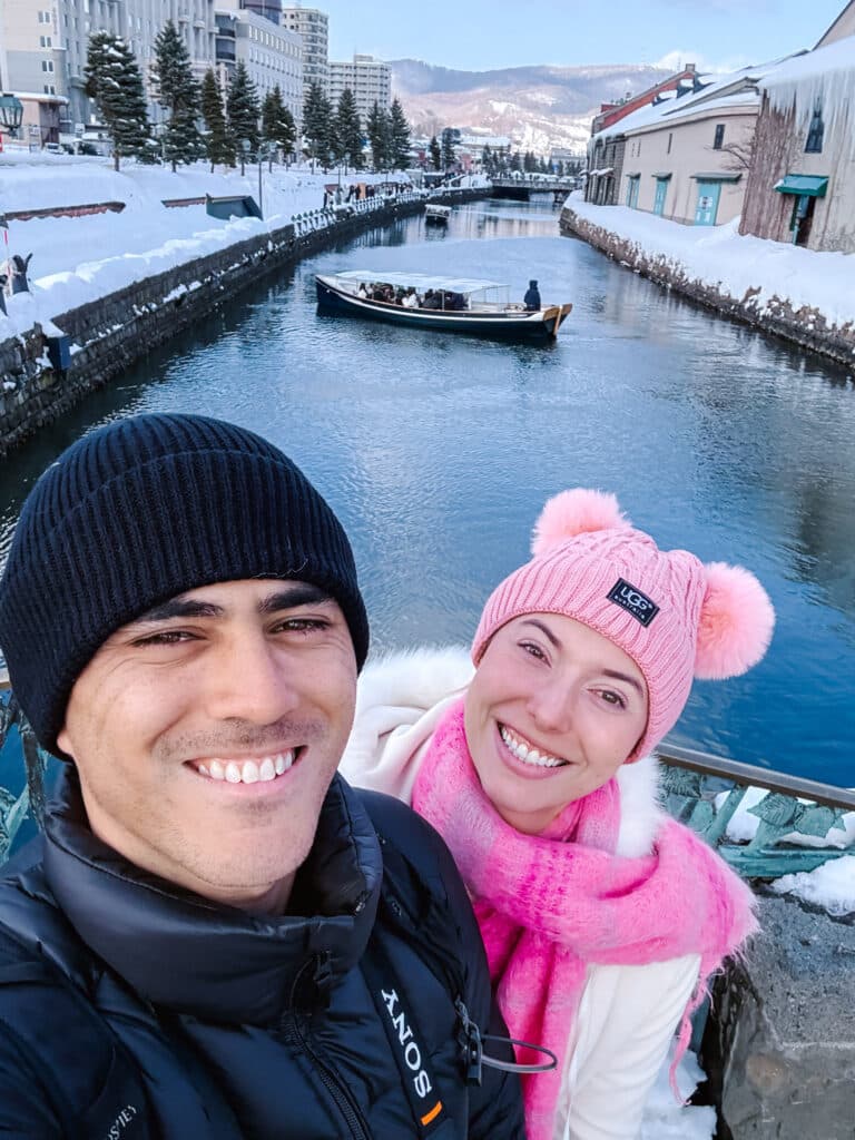 Couple taking a selfie by the Otaru Canal, a cruise boat and snowy warehouses behind them in winter