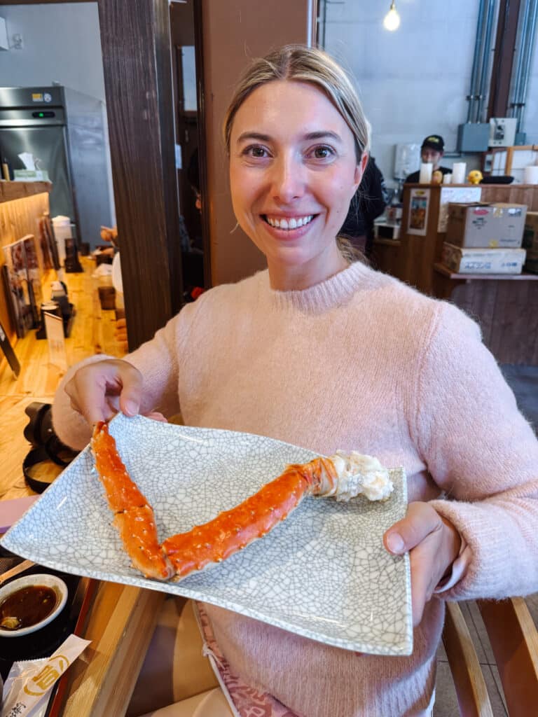 Woman in a pink sweater holding a plate with a long orange king crab leg at Donbei in Otaru