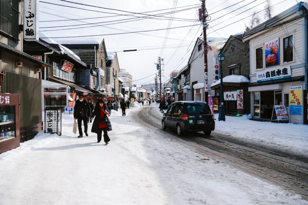 People walking down snow-packed Sakaimachi Street in Otaru, lined with shops and fresh-seafood signs in winter