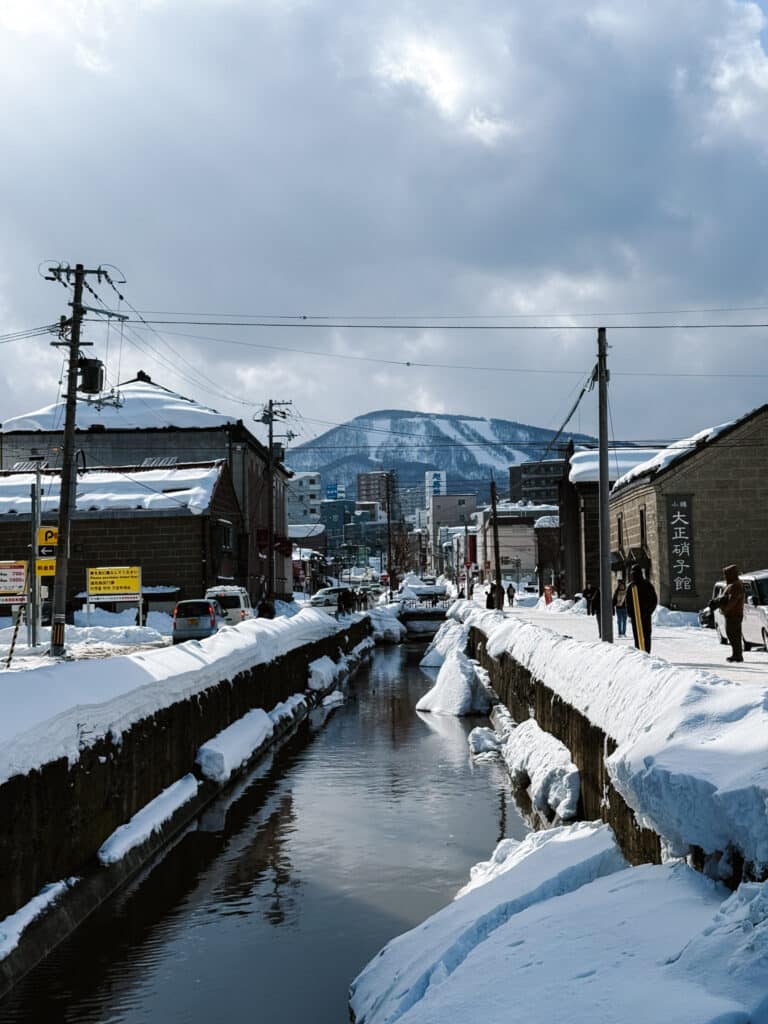 Snow-banked side canal in Otaru reflecting the town, a white mountain rising under a grey winter sky