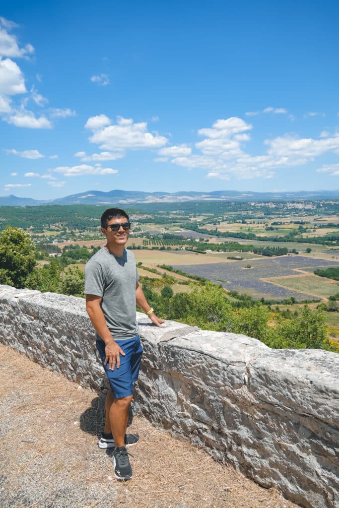 Man in a grey shirt leaning against the stone wall at the Sault village viewpoint with the lavender plateau spread out below