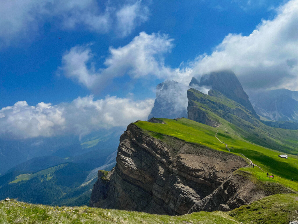 Grassy clifftop viewpoint at Seceda with hikers tracing a path toward cloud-wrapped peaks, Dolomites, Italy
