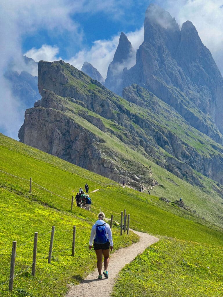 A hiker walking a fenced trail toward the dramatic spires of the Odle peaks at Seceda, Dolomites, Italy