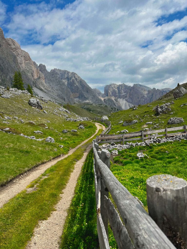 A gravel hiking trail beside a wooden fence winding through the valley toward grey Dolomite peaks, Seceda, Italy