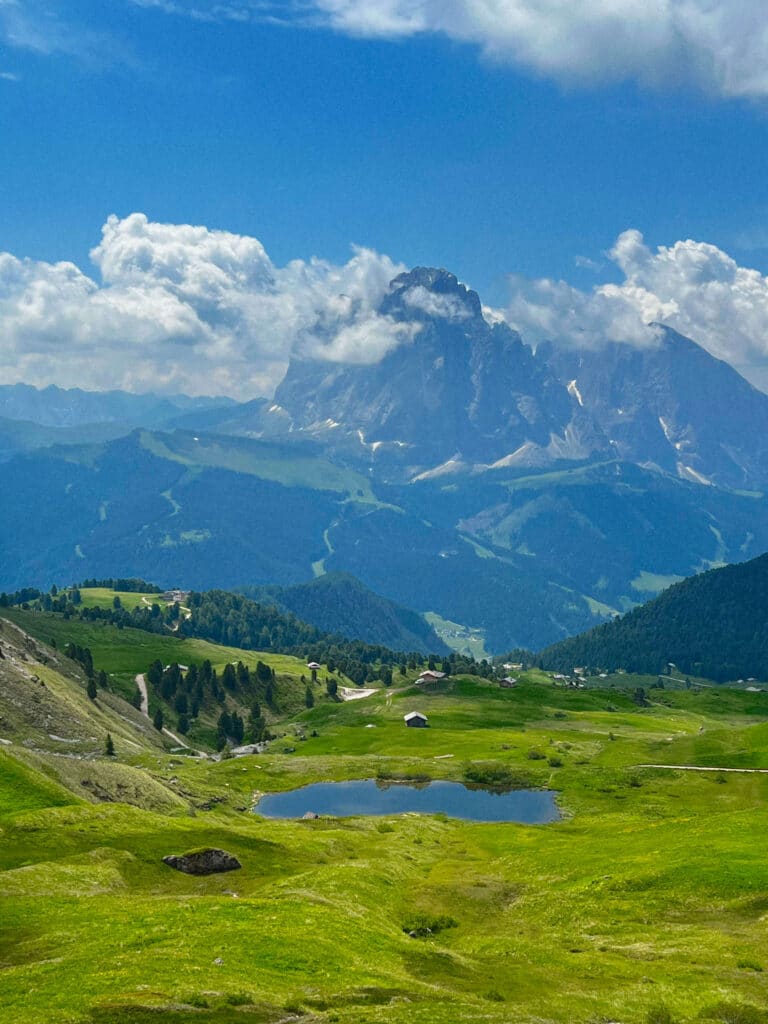 The Sassolungo massif towering over a tarn and scattered mountain huts in a green valley, Seceda, Italy