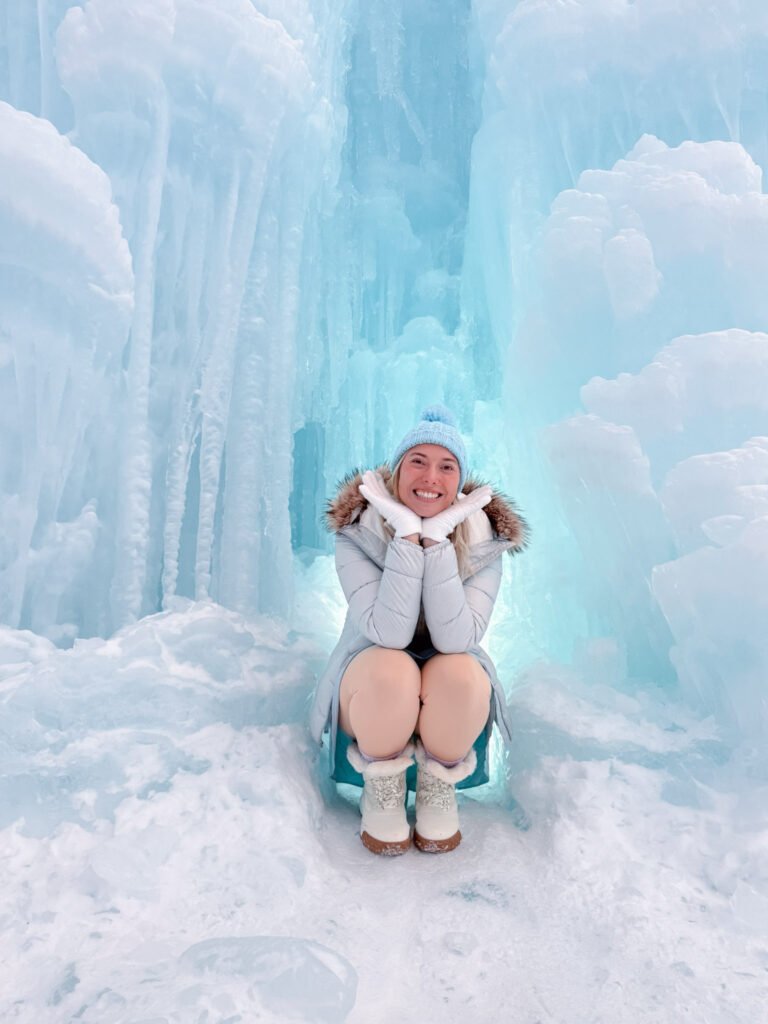 Woman in a silver puffer and pom-pom beanie crouching with hands on cheeks inside a glowing turquoise ice cave at Shikotsu