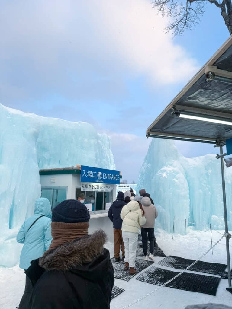 Visitors queuing at the blue "ENTRANCE" ticket booth flanked by sculpted ice walls at the Lake Shikotsu Ice Festival, Hokkaido