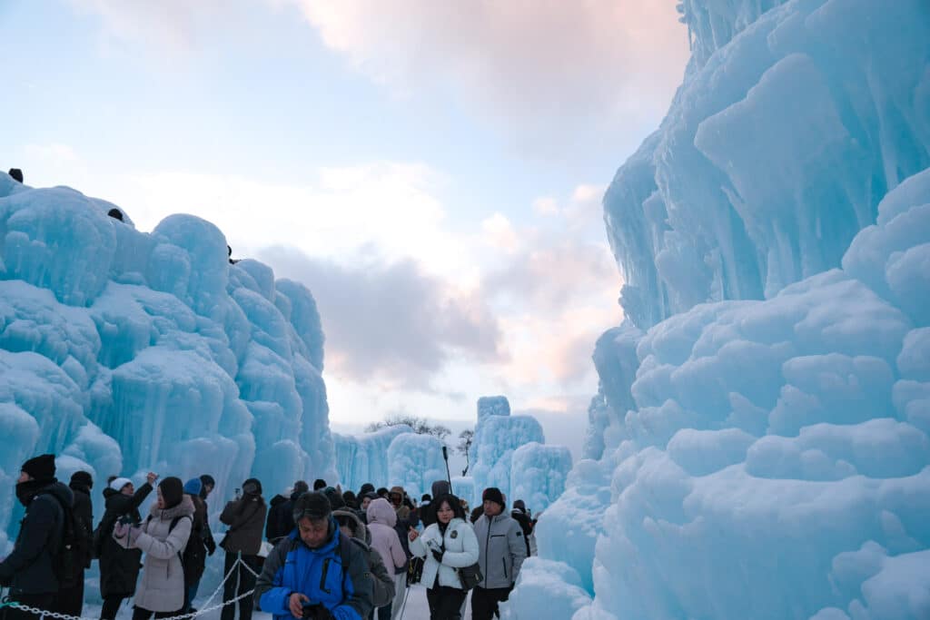Crowds walking through a canyon of towering pale-blue ice sculptures against a soft pink sunset sky at Lake Shikotsu