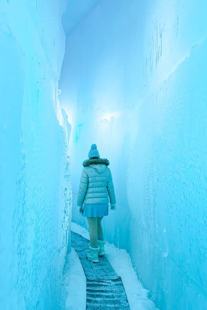 Woman in a pale puffer and pom-pom beanie walking down a narrow glowing blue ice corridor at the Lake Shikotsu Ice Festival