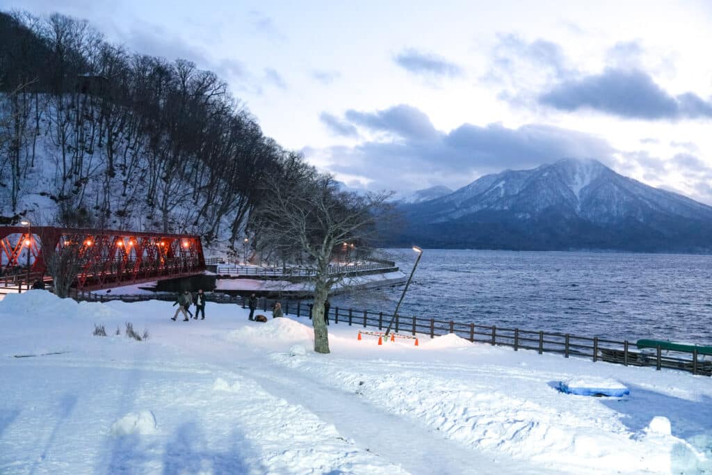 Snowy lakeshore view of the red Yamasen bridge with Mount Eniwa beyond Lake Shikotsu at dusk, Hokkaido in winter.