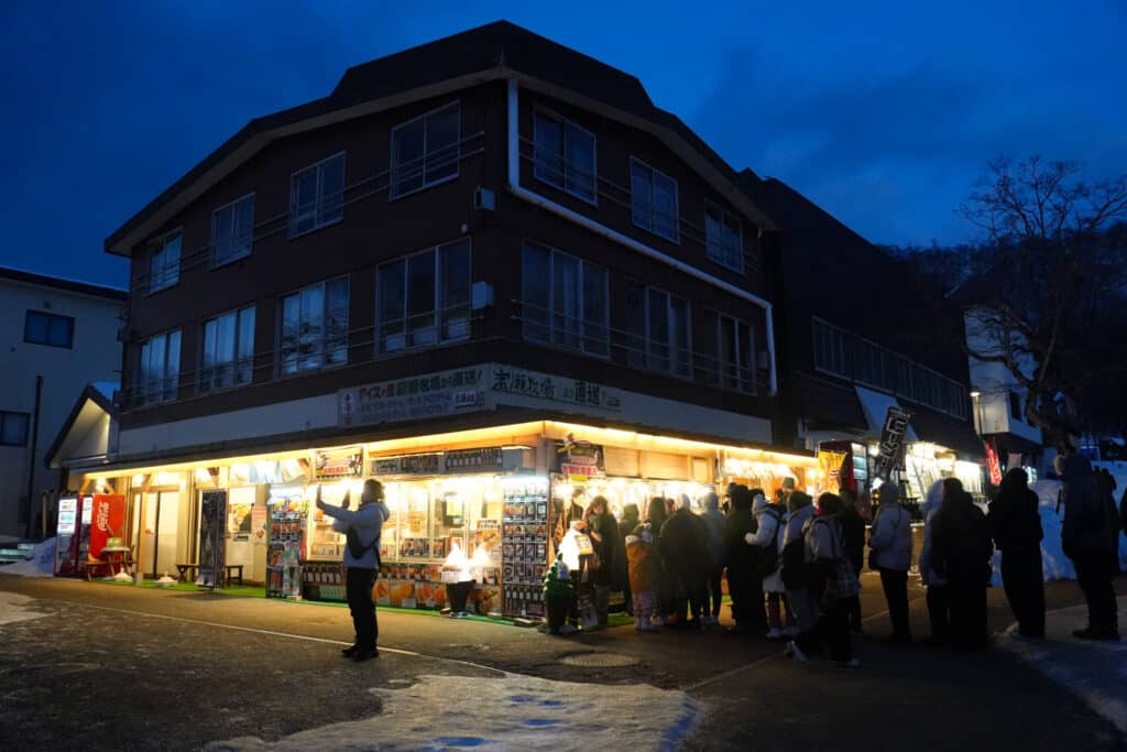 Crowd queuing outside a lit-up Japanese souvenir and snack shop at blue hour near the Lake Shikotsu Ice Festival, Hokkaido