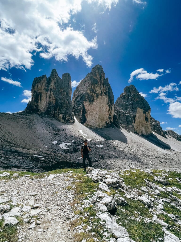 Lone male hiker stands in front of the Tre Cime di Lavaredo north face, dwarfed by the three immense limestone towers