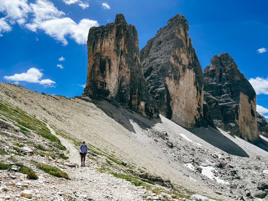 A single hiker on the trail looks tiny against the massive vertical walls of the Tre Cime di Lavaredo north face