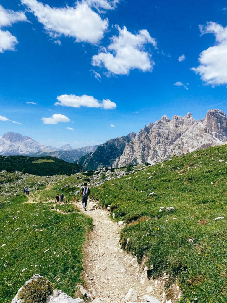 Stony hiking trail winding toward the Tre Cime di Lavaredo through lush green meadows with distant peaks on the horizon