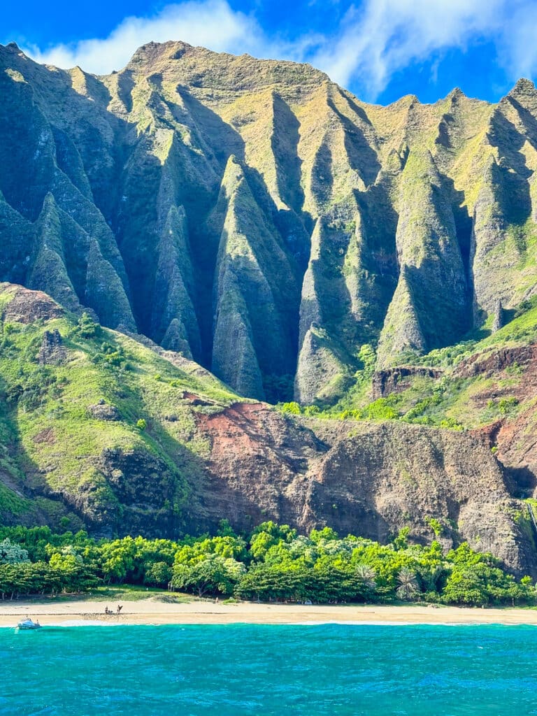 A small tour boat dwarfed by towering green Na Pali cliffs above a sandy beach