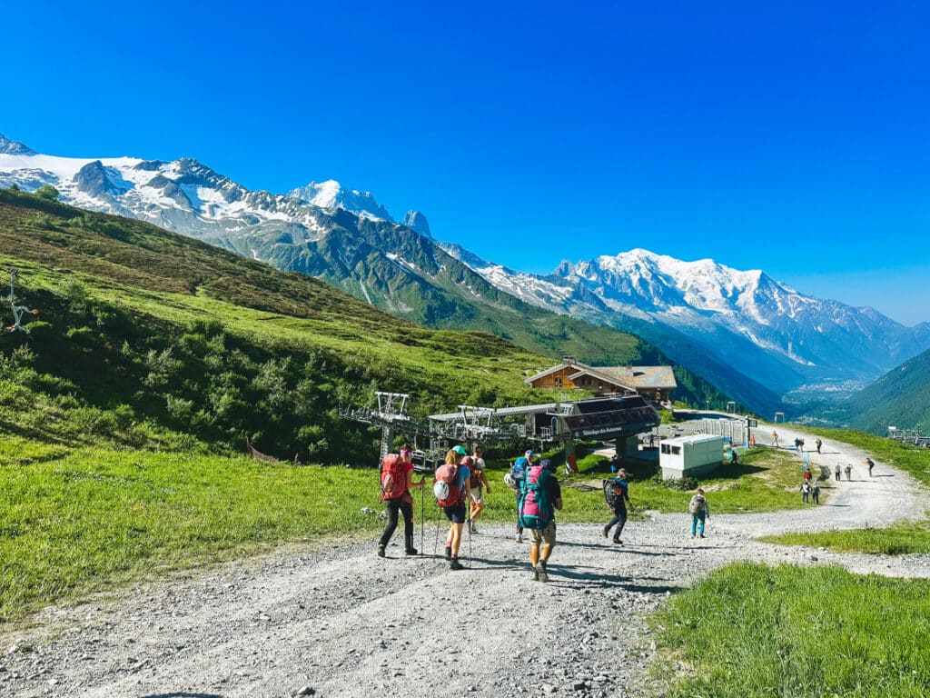 Group of hikers walking a dirt track past a chalet with Mont Blanc behind, Chamonix trailhead