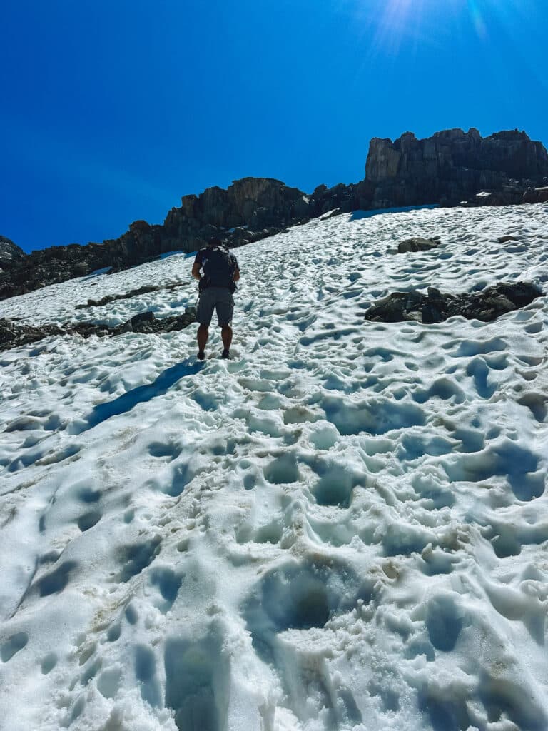 Male hiker climbing a sun-lit snow slope toward rocky crags on the Albert 1er route, French Alps
