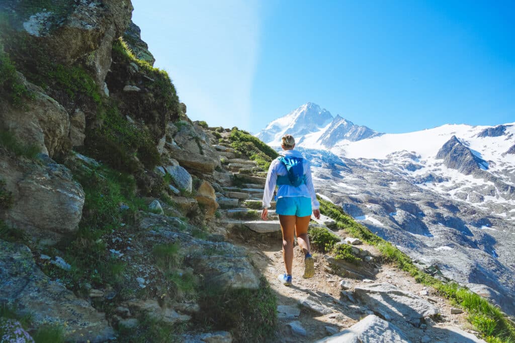 Female hiker climbing stone steps on a cliffside trail toward a snowy peak, Albert 1er hike, Chamonix