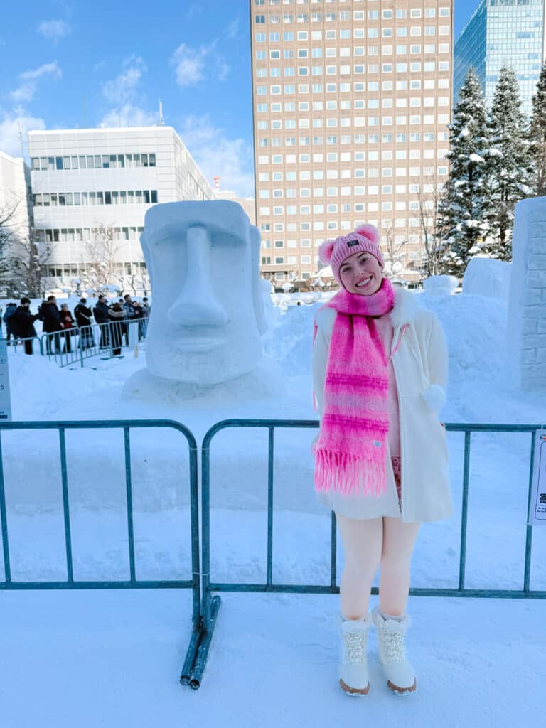 Visitor posing beside a Moai head snow sculpture under blue sky at the Sapporo Snow Festival, Hokkaido, Japan