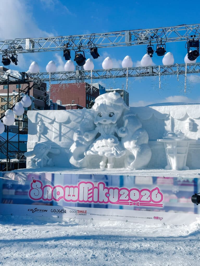 Snow Miku 2026 snow sculpture of the white-and-aqua Hatsune Miku character above a branded festival banner at Odori Park, Sapporo, Japan