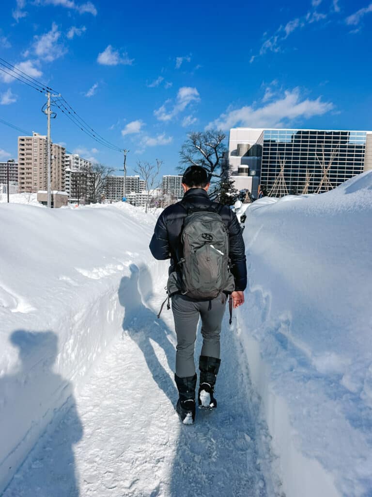 Traveller walking a cleared path between tall snowbanks near Odori park under blue sky in wintry Sapporo, Hokkaido, Japan