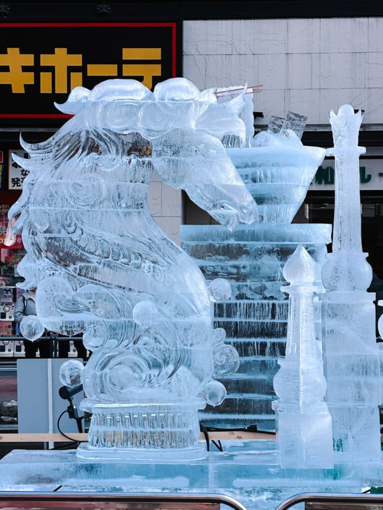 Translucent ice sculpture of a horse and chess-piece motifs lit against Susukino's neon storefronts at Sapporo Snow Festival, Japan