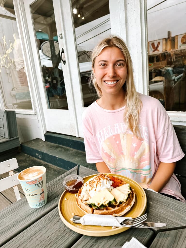 Woman smiling with a Java Kai shark bite latte and a tropical waffle at the outdoor tables of the Kapaa cafe on Kauai