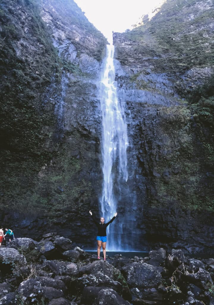 Hiker with arms raised at the base of towering Hanakāpīʻai Falls near Hāʻena State Park, Kauaʻi