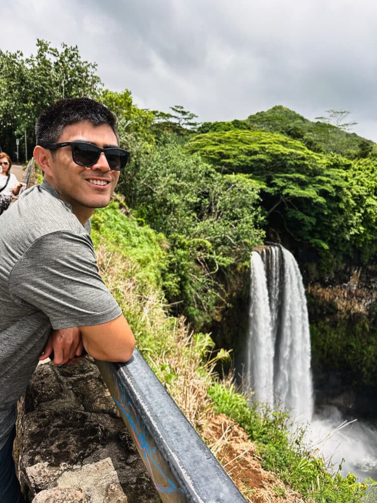 Traveler leaning on the railing at the Wailua Falls roadside lookout with the waterfall behind, Kauai, Hawaii