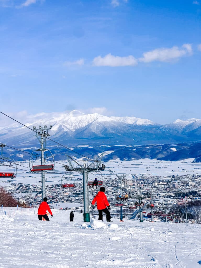 Two snowboarders in red jackets at the Furano Ski Resort with the snow-capped Tokachi mountains and blue skies beyond